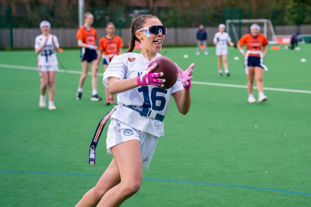 A young athlete catches a football during a flag football game run by Edgerush Flag Football, with teammates watching in the background on a sports field.