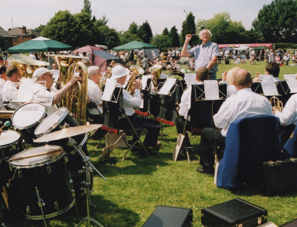 A brass band performing outdoors at Parbold Village Show, with a conductor leading the musicians as they play to an audience on a sunny field.