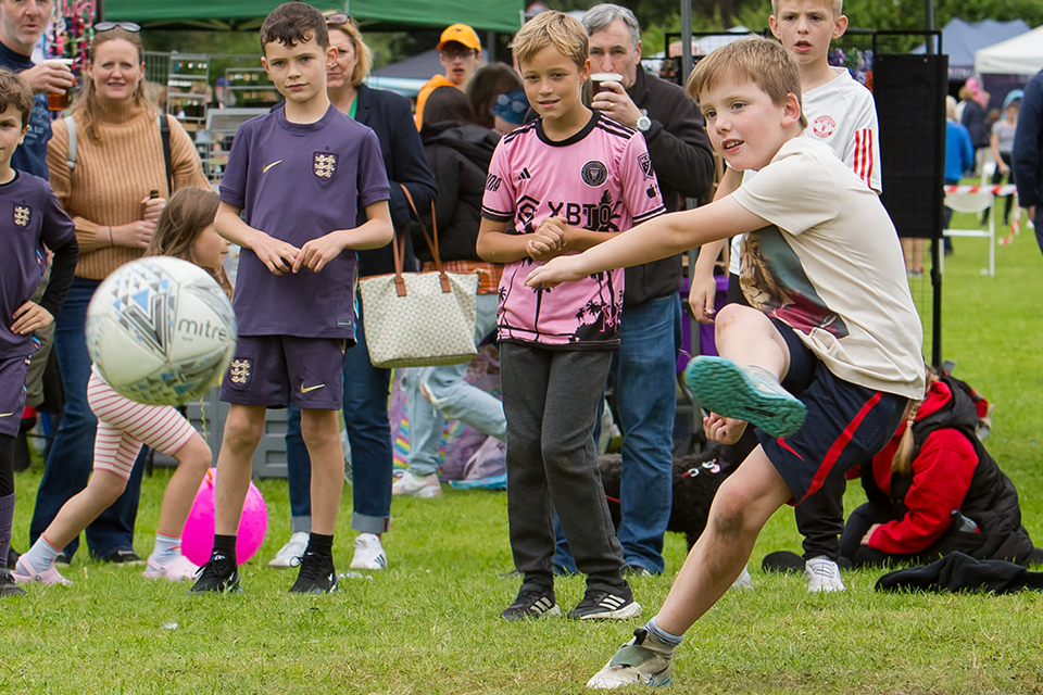 Photo of young boy kicking a ball towards the camera, a crowd of young kids looks on behind him. 