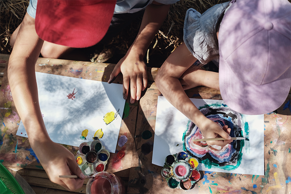 Photograph taken from above of two children doing craft and painting activities outdoors on a wooden table. 