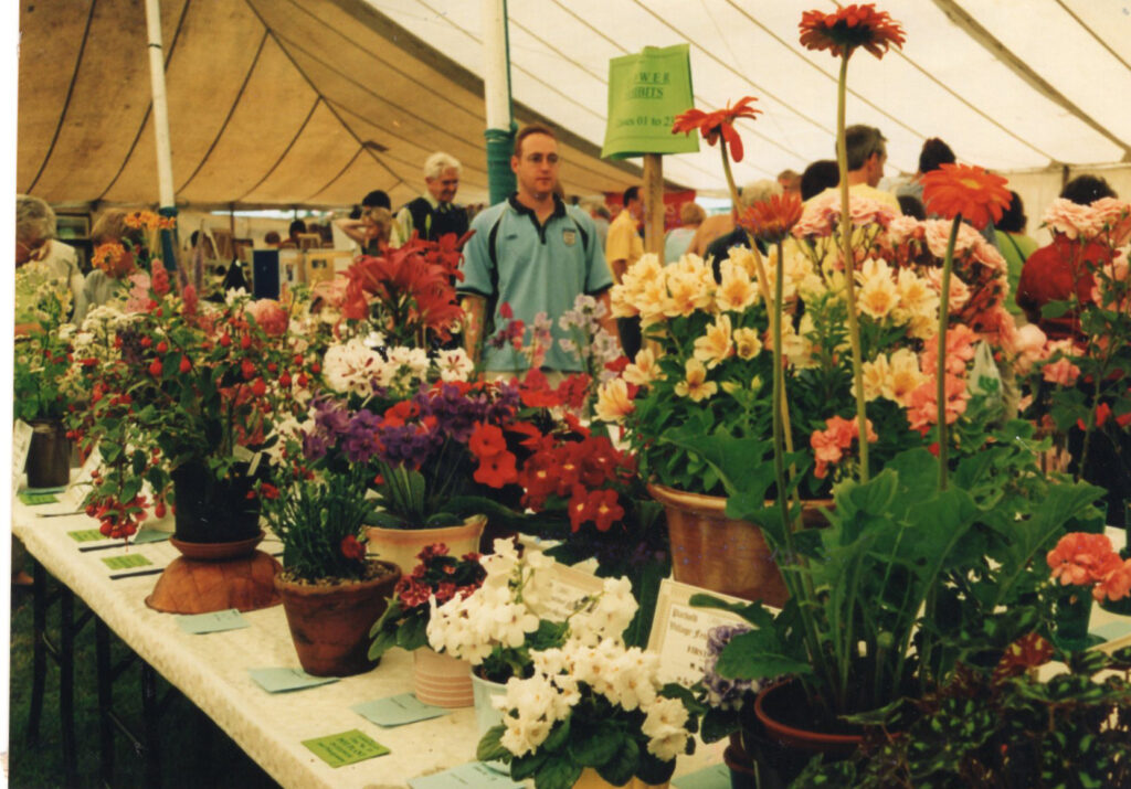 Colourful potted flower arrangements on display in the Show Tent at Parbold Village Show, with visitors admiring the entries along long tables under a large marquee.