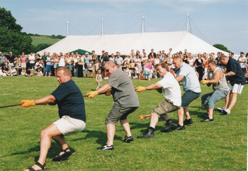 A team of men competing in a tug of war at Parbold Village Show, pulling a rope with determination as a large crowd watches in front of a white marquee.
