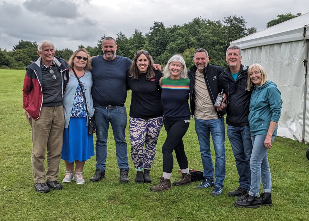 A group of smiling volunteers standing arm in arm on the show field at Parbold Village Show, posing in front of a white marquee on a cloudy day.