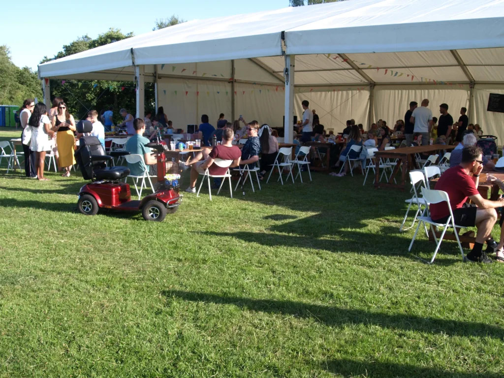 Crowds of families and friends enjoying food and drinks under a large marquee at the Parbold Village Show Friday Night Disco 2025, with summer sun and bunting overhead.