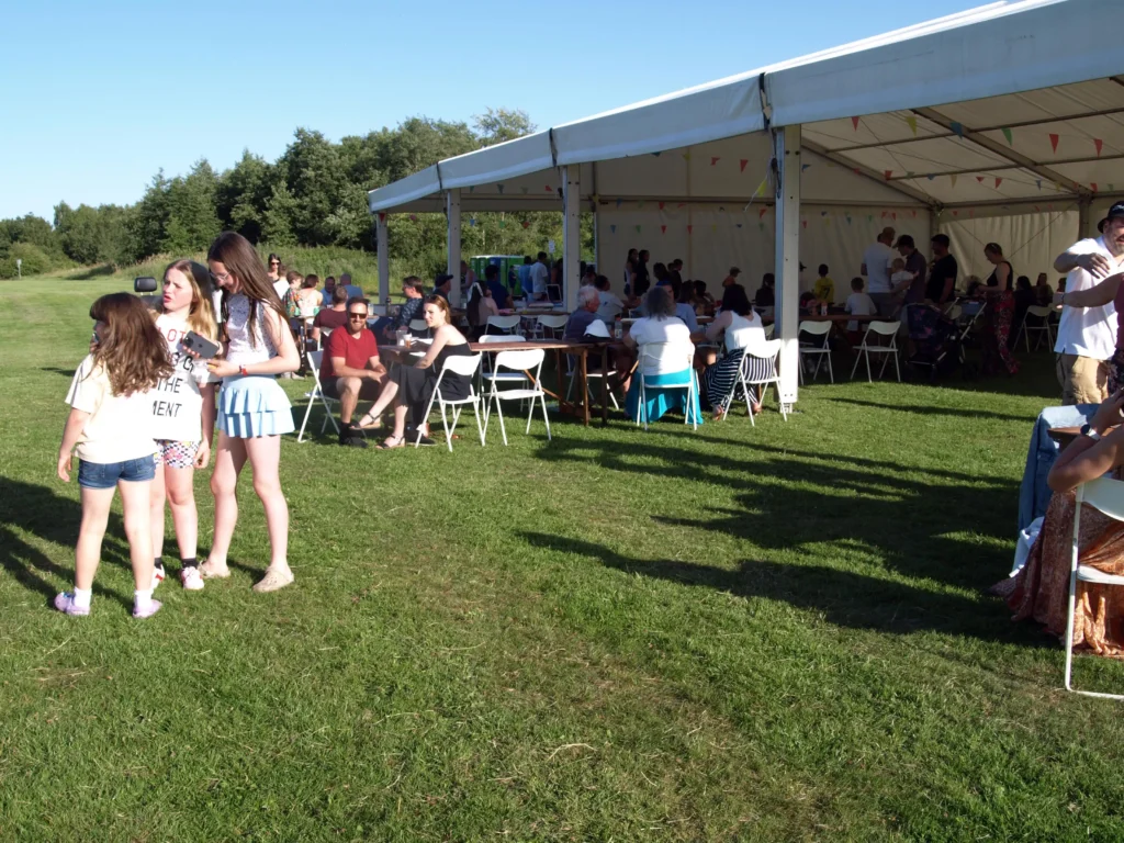 Groups of people sitting and chatting outside the marquee at the Parbold Village Show Friday Night Disco 2025, with children playing on the field nearby.