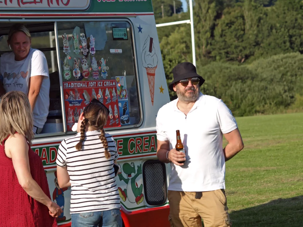 Man drinking beer while families queue for ice cream from a colourful van at the Parbold Village Show Friday Night Disco 2025.