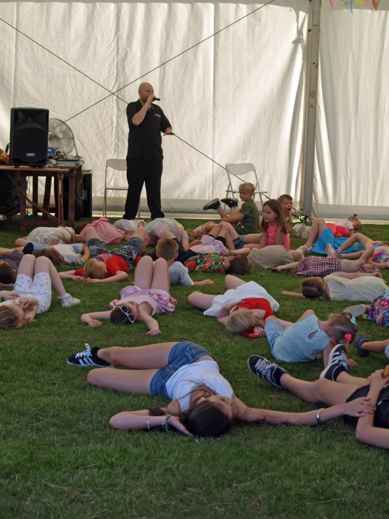 Performer speaking into a microphone while children take part in a group game inside the marquee at the Parbold Village Show Friday Night Disco 2025.