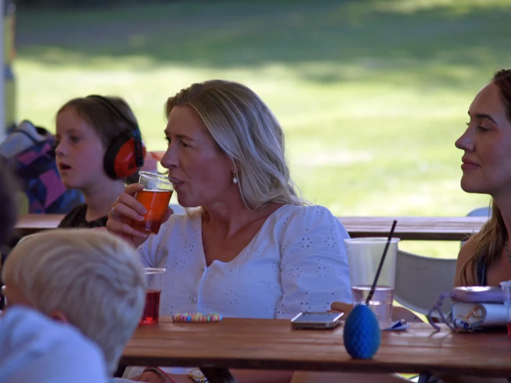 Woman drinking from a plastic cup at a picnic table, while children and families enjoy the atmosphere at the Parbold Village Show Friday Night Disco 2025.