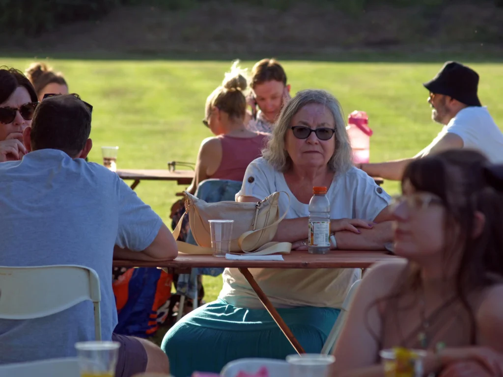 Older woman in glasses seated at a picnic bench during the Parbold Village Show Friday Night Disco 2025, with others relaxing nearby in the evening sunshine.