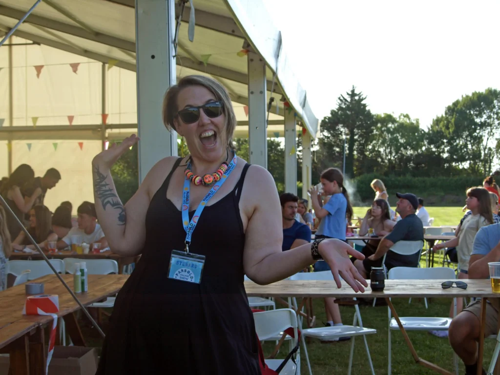 Parbold Village Show steward posing happily for the camera outside the marquee at the Friday Night Disco 2025, with festivalgoers seated in the background.