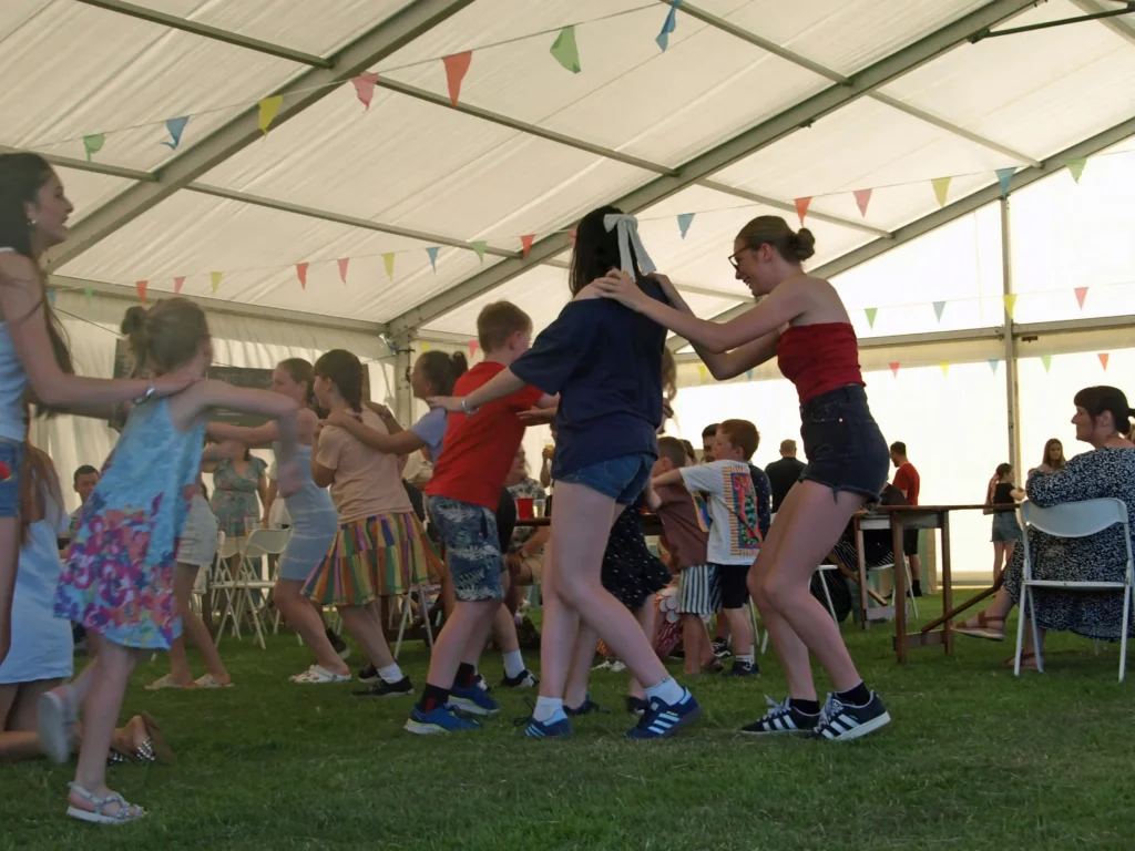 Children dancing in a conga line under the marquee at the Parbold Village Show Friday Night Disco 2025, with colourful bunting overhead.