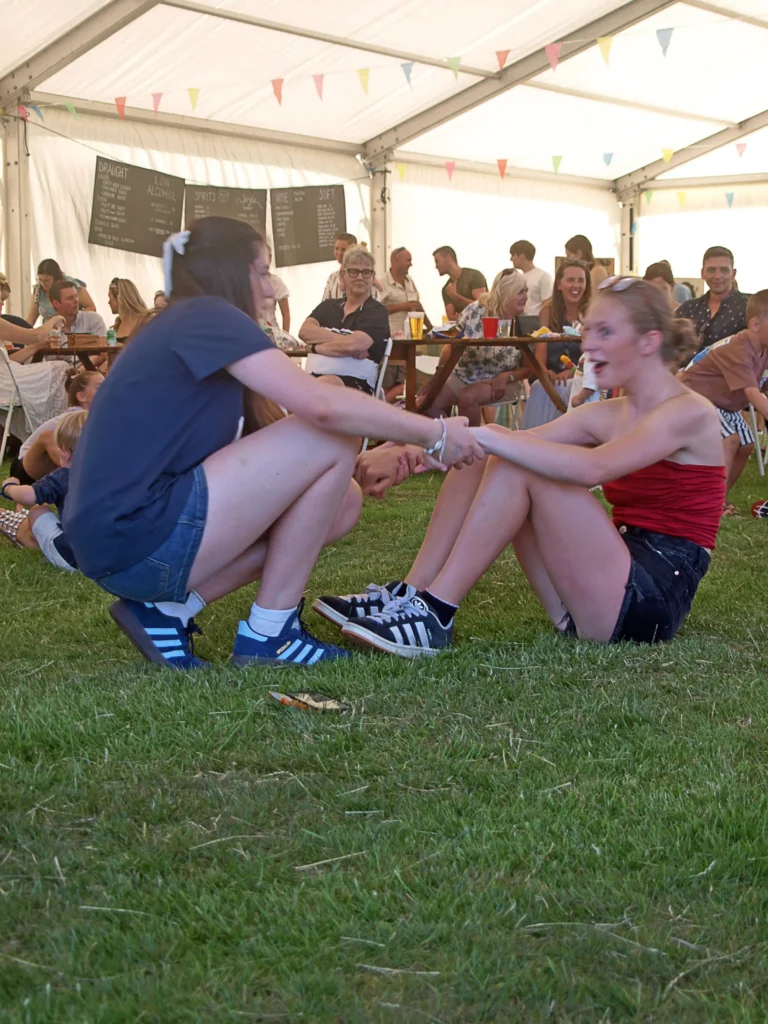 Two older children taking part in a seated tug-style game inside the marquee at the Parbold Village Show Friday Night Disco 2025, surrounded by smiling guests.