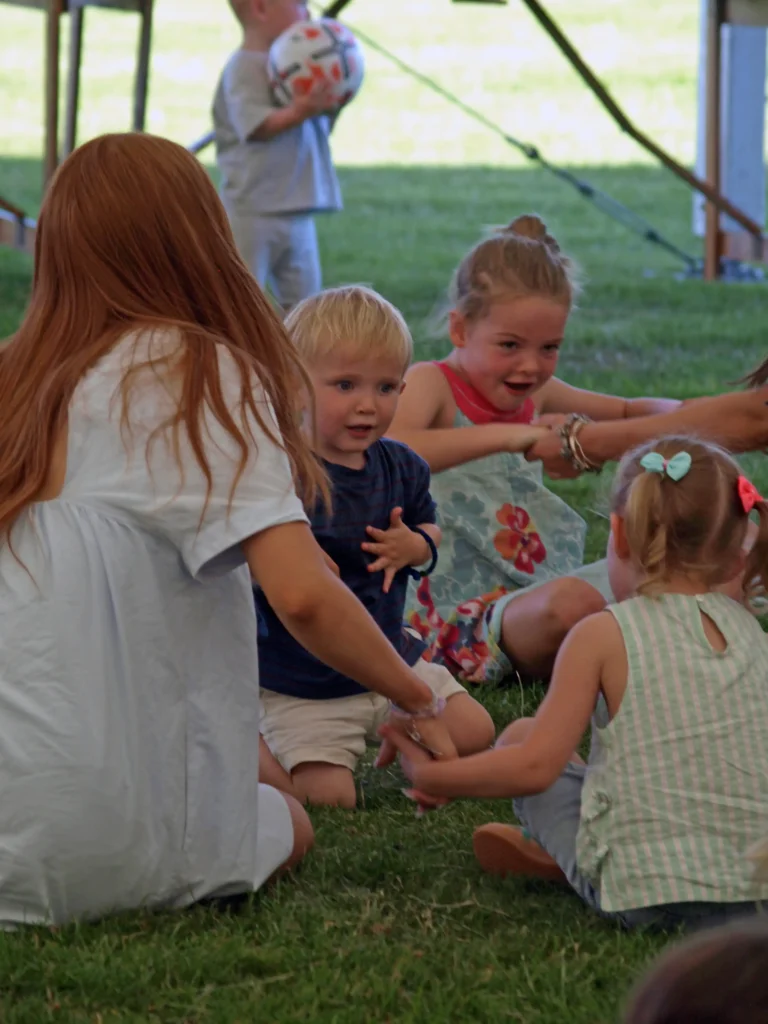 Group of young children sitting on the grass, playing a circle game with an adult during the Parbold Village Show Friday Night Disco 2025.