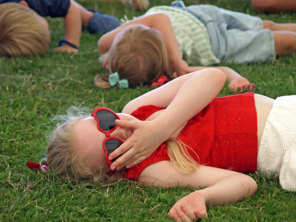 Young girl in red sunglasses lying on the grass pretending to sleep during a game at the Parbold Village Show Friday Night Disco 2025.