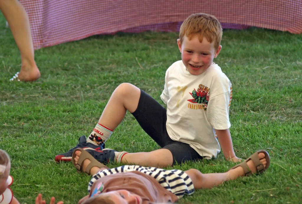 Laughing young boy sitting on the grass under the marquee at the Parbold Village Show Friday Night Disco 2025, after taking part in a group game.