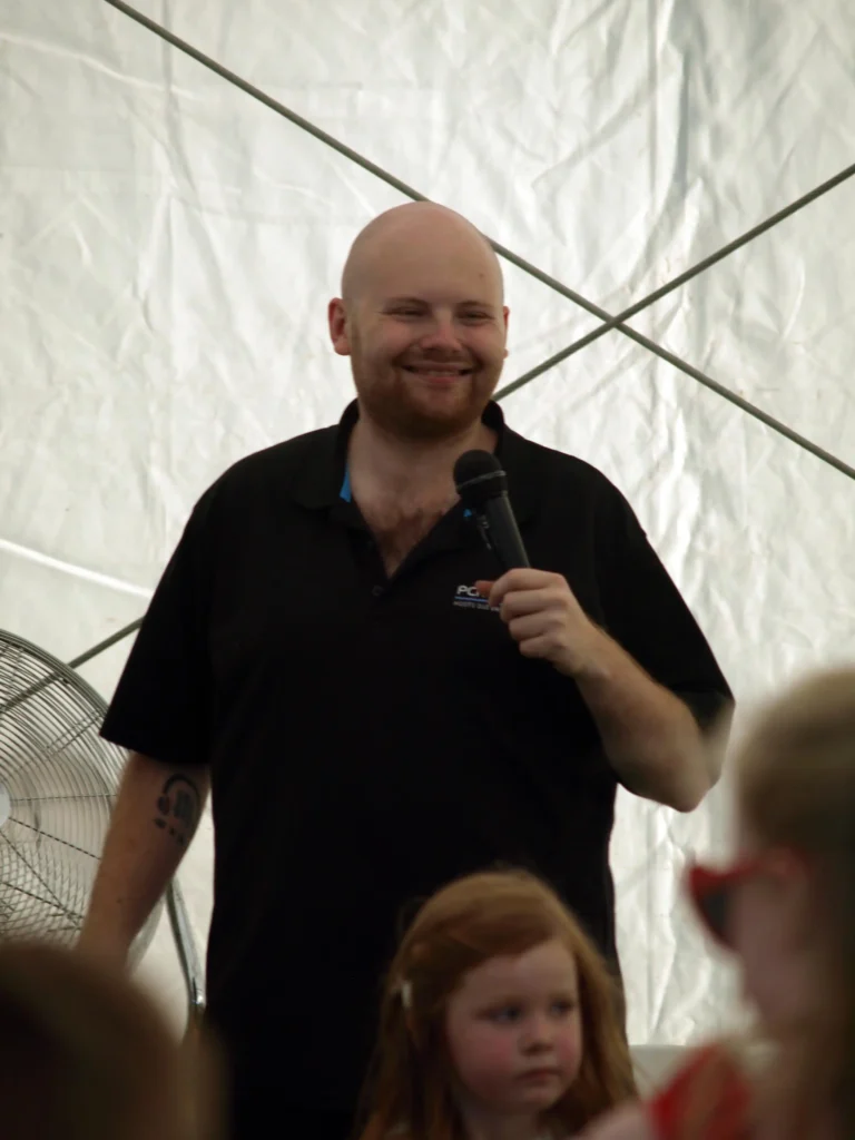 Smiling entertainer holding a microphone while leading children’s games under the marquee at the Parbold Village Show Friday Night Disco 2025.