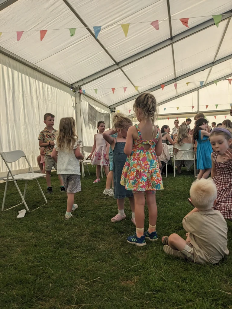 Children preparing for a musical chairs-style game inside the marquee at the Parbold Village Show Friday Night Disco 2025, with colourful bunting above.