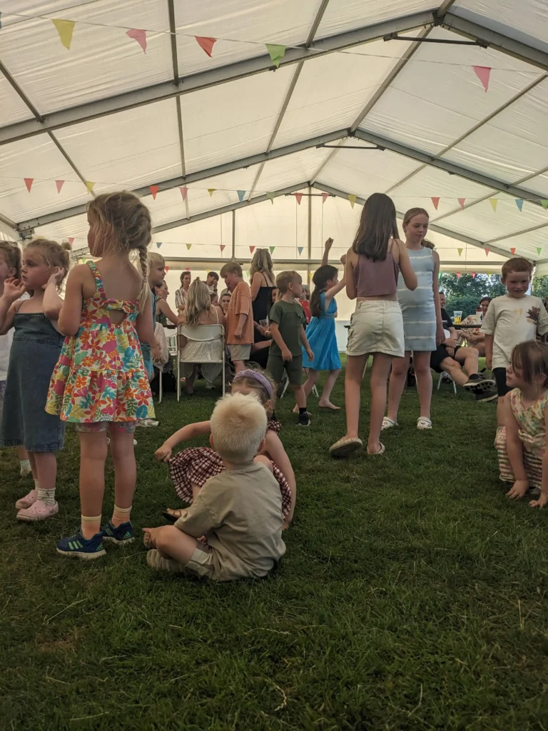 Kids gathering and chatting ahead of a party game at the Parbold Village Show Friday Night Disco 2025, with a large crowd seated in the background.