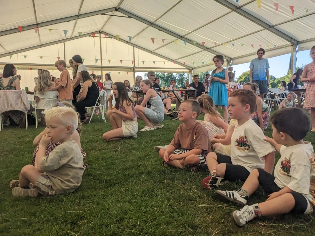 Front row of children seated on the grass watching a performance at the Parbold Village Show Friday Night Disco 2025, with families behind.