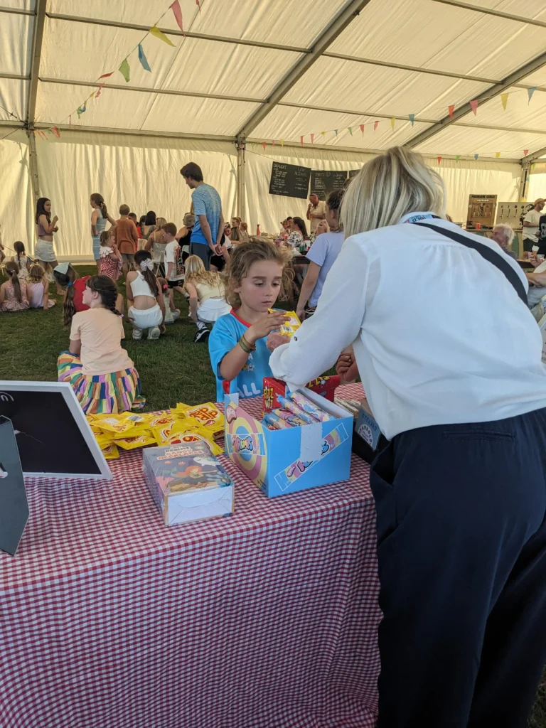 Child choosing sweets at the tuck shop table under the marquee at the Parbold Village Show Friday Night Disco 2025, surrounded by seated families.