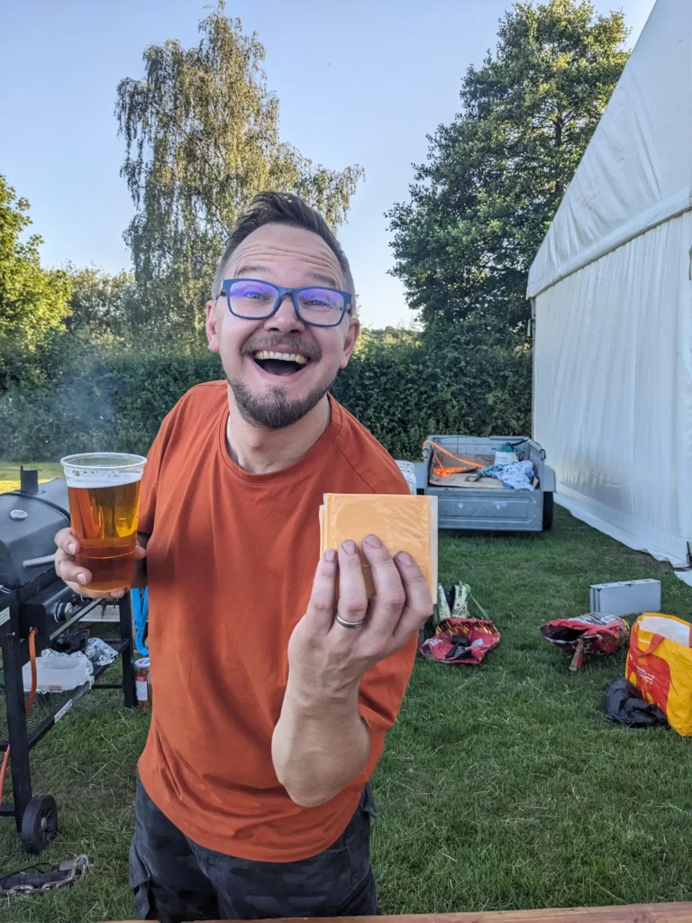 A man in an orange t-shirt grinning widely while holding a pint of beer in one hand and a stack of cheese slices in the other, standing next to a barbecue.