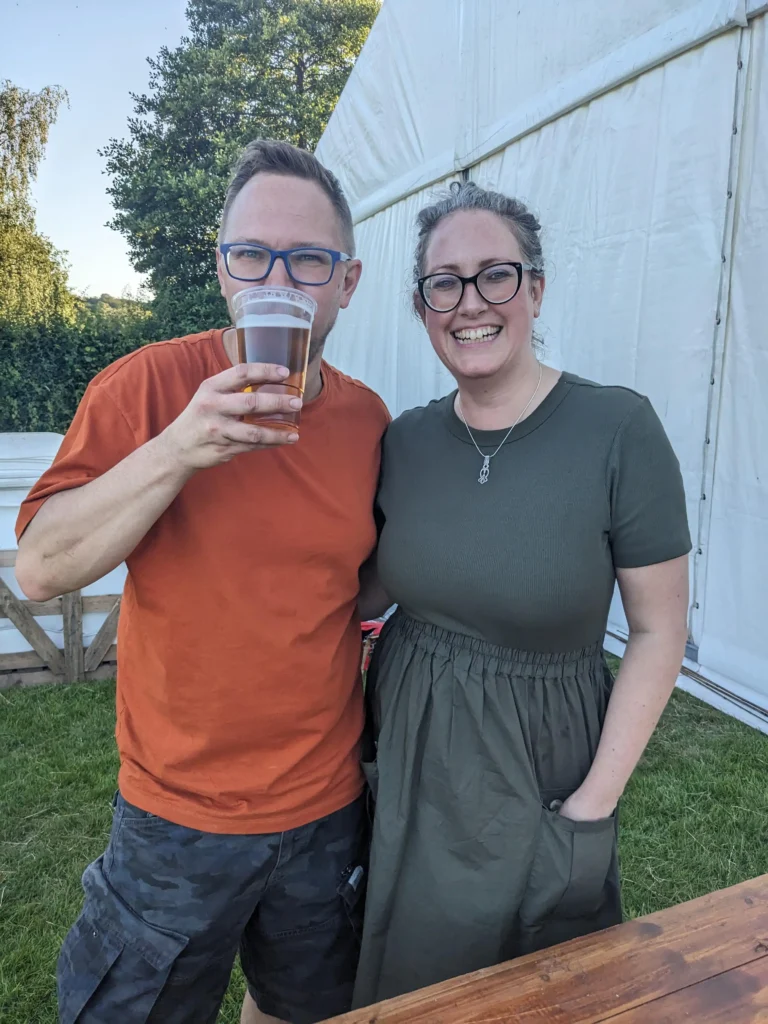 A smiling man and woman standing together outside a marquee; the man holds a pint of beer and the woman wears glasses and a green dress.