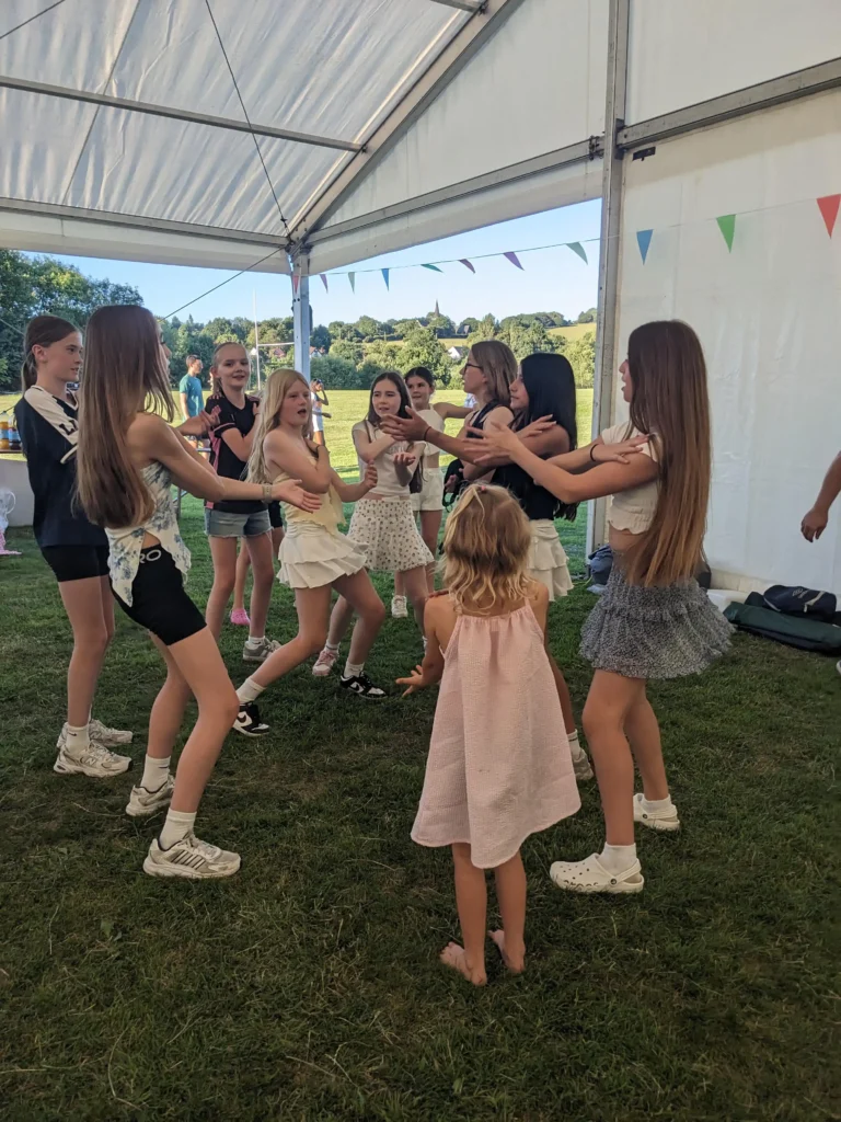 Teen and pre-teen girls dancing in a circle under a marquee, with a barefoot younger girl in a pink dress joining in from the middle.